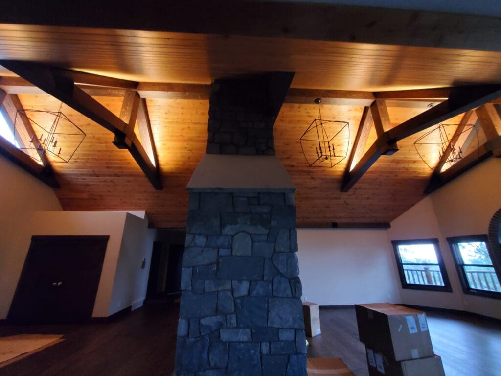 Vaulted ceiling with exposed timber beams and custom woodwork, featuring modern chandeliers and a floor-to-ceiling stone fireplace, remodeled by JG Carpentry in Spearfish, SD.