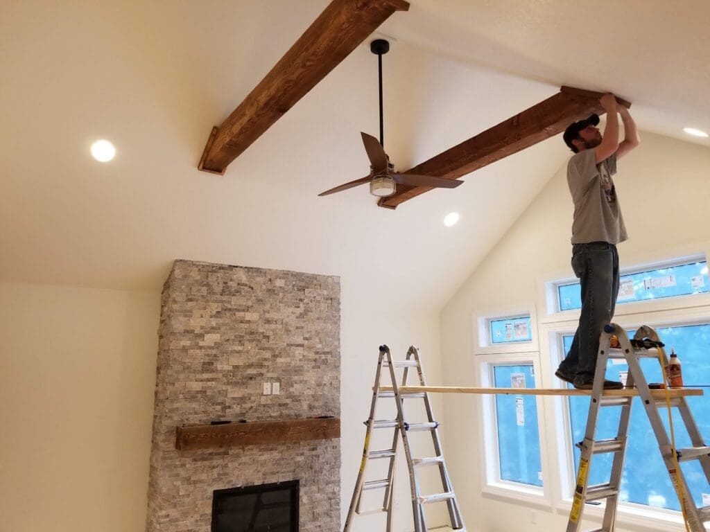 JG Carpentry carpenter installing rustic wood beams on a vaulted living room ceiling in Spearfish, SD, with a floor-to-ceiling stacked stone fireplace and wood mantel below.