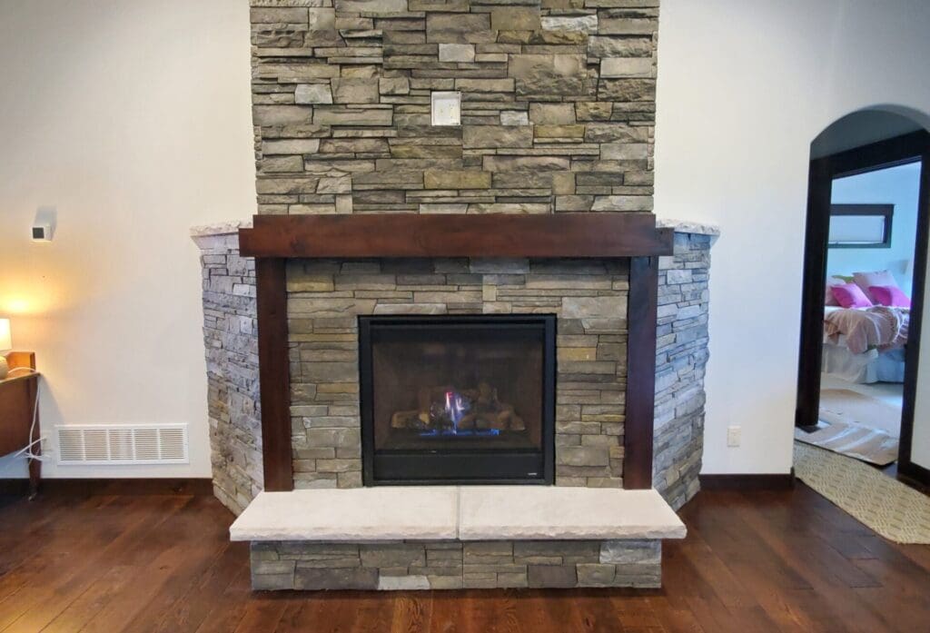 Custom corner fireplace by JG Carpentry in Spearfish, SD: floor-to-ceiling stacked stone, dark-stained wood mantel and posts, raised stone hearth, and a gas fireplace insert.
