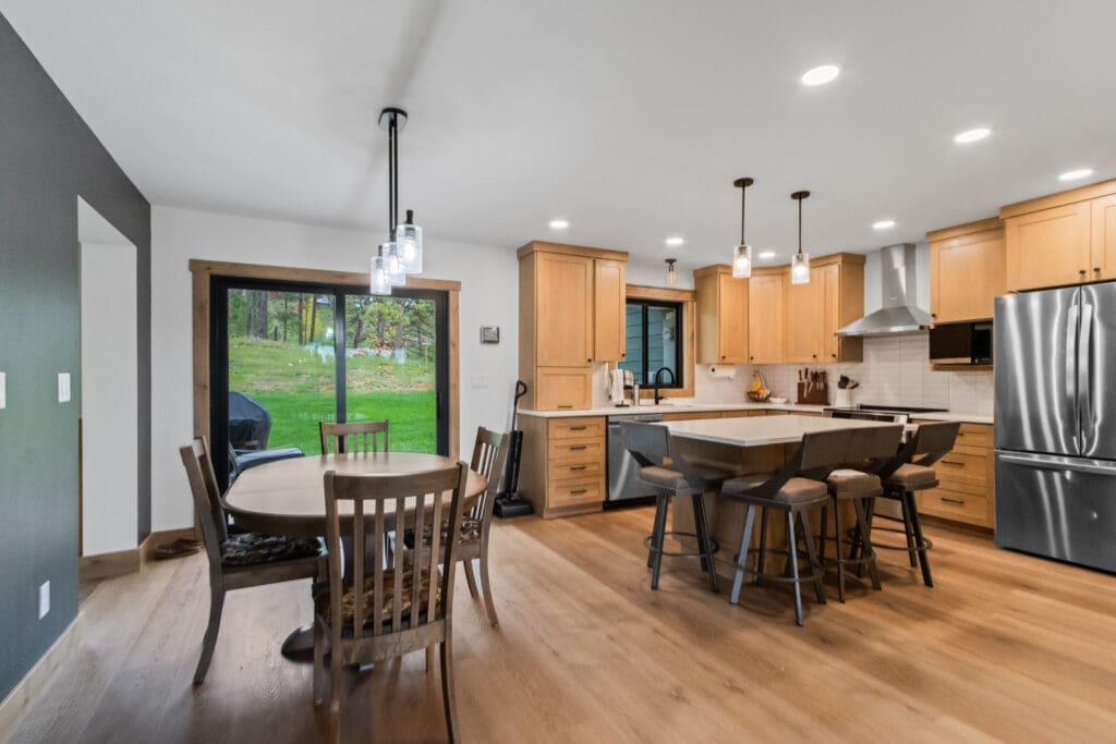 Whole-home remodel in Spearfish, SD by JG Carpentry: open kitchen and dining area with maple shaker cabinets, white quartz island with seating, glass pendant lights, and wood-look plank flooring.