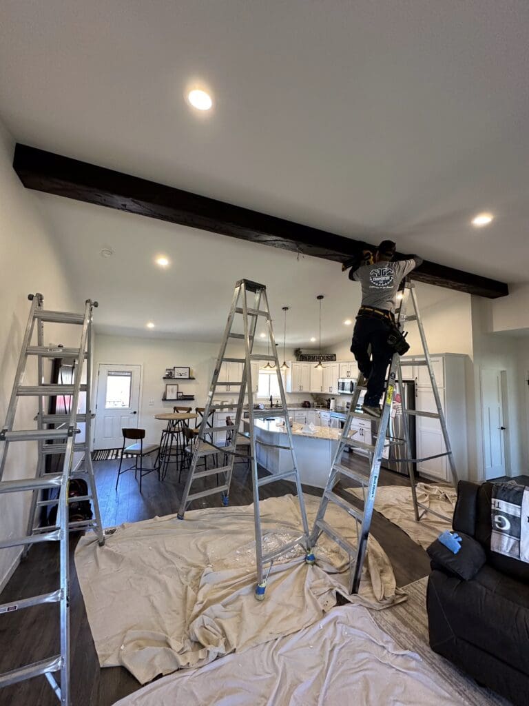 JG Carpentry team installing a dark wood ceiling beam during a kitchen remodel in Spearfish, SD. The scene shows multiple ladders, protective floor coverings, and a modern kitchen layout with pendant lighting and white cabinetry in progress.